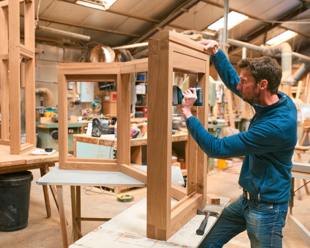 Carpenter In Workshop Using Drill To Attach Piece Of Wood To Window Frame